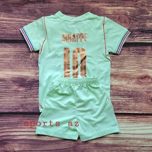 May include: A light green short-sleeved jersey and matching shorts set. The jersey features the name MBAPPE and the number 10 in metallic copper lettering on the back. The sleeves have blue, white, and red striped trim. The text sports az is visible.
