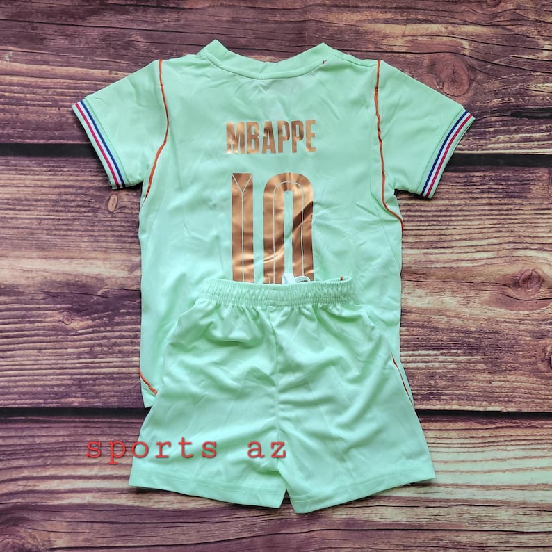 May include: A light green short-sleeved jersey and matching shorts set. The jersey features the name MBAPPE and the number 10 in metallic copper lettering on the back. The sleeves have blue, white, and red striped trim. The text sports az is visible.