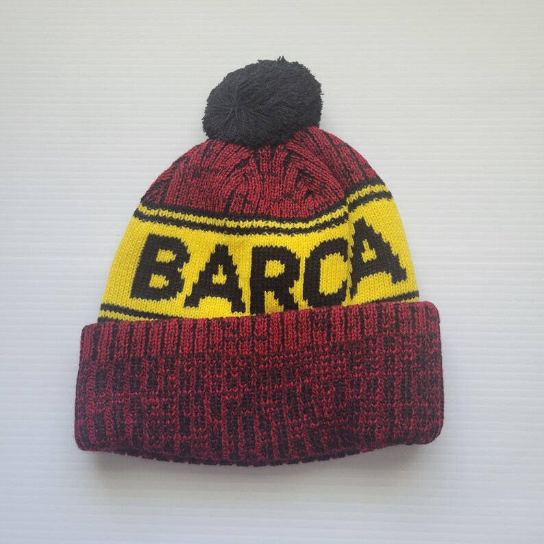May include: A red and black knit beanie with a yellow band and the word "BARCA" in black letters. The beanie has a black pom-pom on top.
