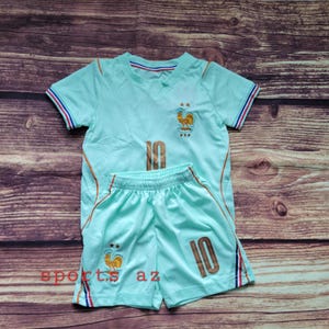 May include: A light mint green short-sleeved jersey and matching shorts set featuring the number 10 in gold, a gold rooster emblem with two stars above it, and red, white, and blue striped trim on the sleeves and shorts. The text sports az is visible.