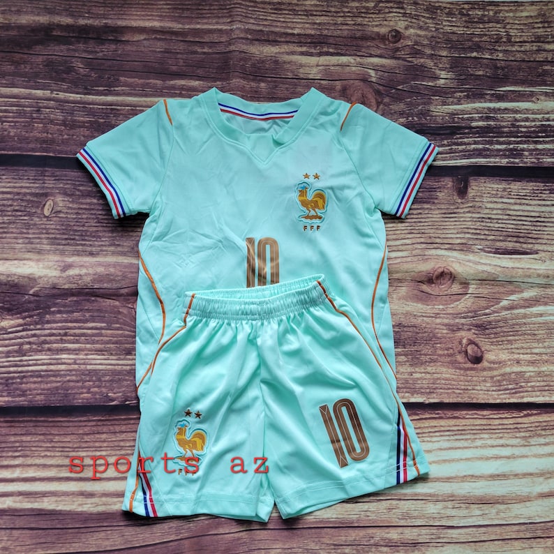 May include: A light mint green short-sleeved jersey and matching shorts set featuring the number 10 in gold, a gold rooster emblem with two stars above it, and red, white, and blue striped trim on the sleeves and shorts. The text sports az is visible.