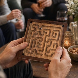 May include: A square wooden maze puzzle game featuring a light-colored wood interior with carved pathways and a dark wood frame. A small silver metal ball is positioned within the maze tracks, which are covered by a clear protective panel secured by screws.