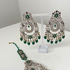 May include: An Indian bridal jewellery set in green and gold, featuring a tikka, earrings and a necklace. The jewellery is decorated with small mirrors and green stones.