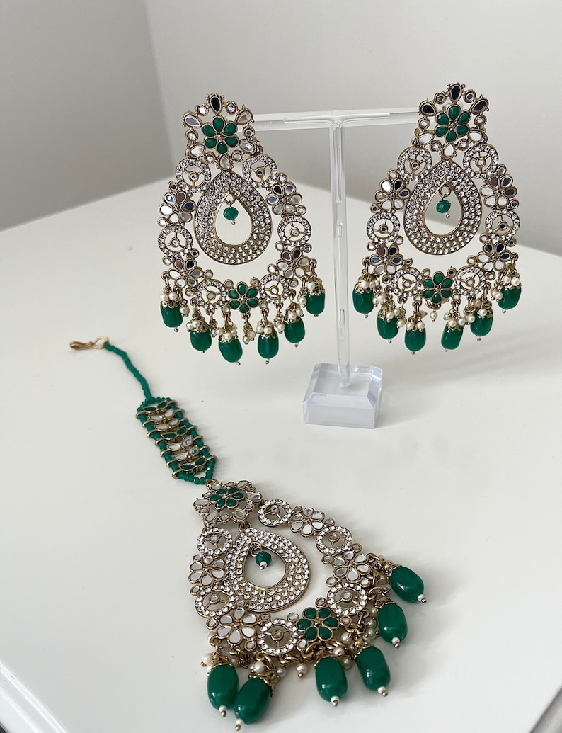 May include: An Indian bridal jewellery set in green and gold, featuring a tikka, earrings and a necklace. The jewellery is decorated with small mirrors and green stones.