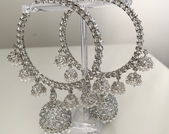 May include: A pair of silver hoop earrings with dangling charms. The charms are round and have a white stone design. The earrings are on a white stand.
