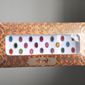Può includere: Una scatola di 18 bindis adesivi di forma ovale e di colori vivaci. La scatola è dorata e riporta il testo "Exclusive Wedding Bindi" e "vivah DESIGNER BINDI".