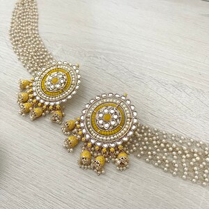 May include: A pair of yellow and gold earrings with pearl accents. The earrings feature a round design with a central yellow flower and multiple layers of pearls. The earrings are attached to a chain of pearls.