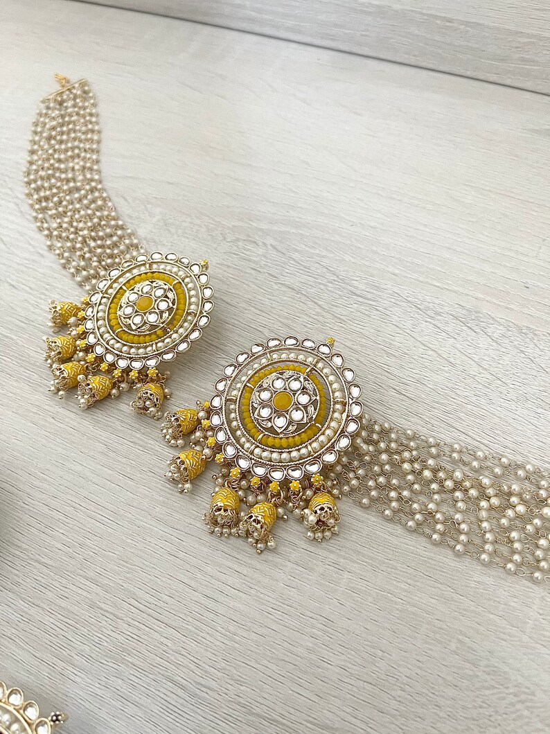 May include: A pair of yellow and gold earrings with pearl accents. The earrings feature a round design with a central yellow flower and multiple layers of pearls. The earrings are attached to a chain of pearls.