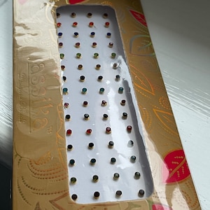 May include: A sheet of colourful rhinestones in a clear plastic pack with a gold background. The pack has the text "Assa" and "Designer Bindi" printed on it.