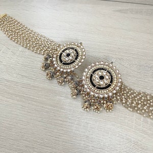 May include: A pair of gold earrings with black and white stones and pearl accents. The earrings have a round design with a central black stone surrounded by smaller white stones. The earrings are attached to a chain of pearls.