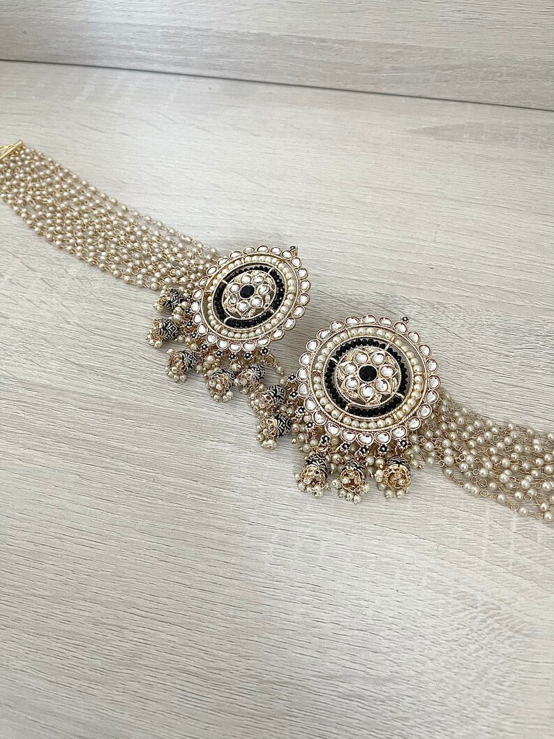 May include: A pair of gold earrings with black and white stones and pearl accents. The earrings have a round design with a central black stone surrounded by smaller white stones. The earrings are attached to a chain of pearls.