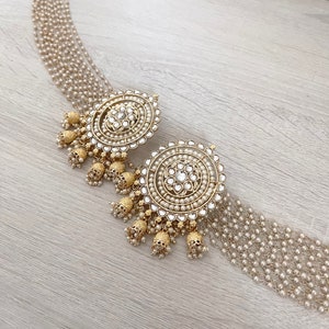May include: A pair of gold and white round earrings with pearl accents and dangling charms. The earrings are on a wooden surface.