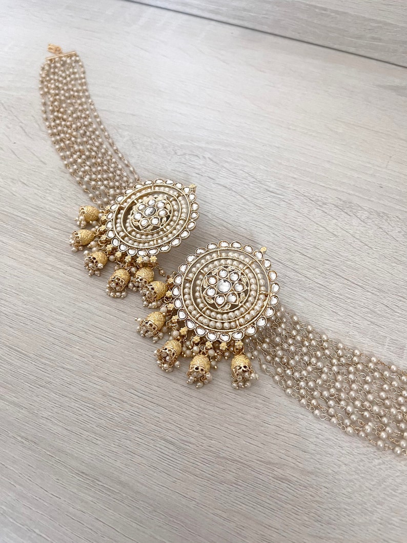 May include: A pair of gold and white round earrings with pearl accents and dangling charms. The earrings are on a wooden surface.