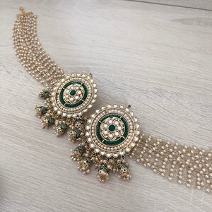 May include: A pair of gold earrings with green and white stones. The earrings have a round shape and are decorated with intricate details. The earrings are hanging from a chain of pearls.