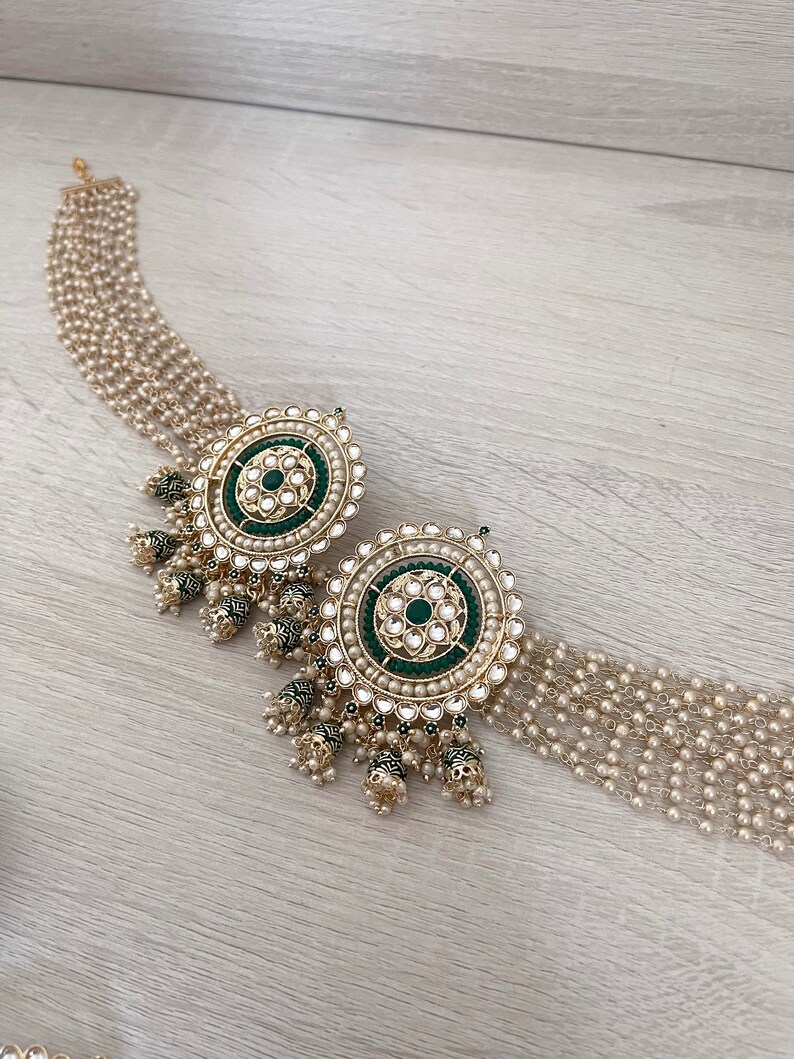 May include: A pair of gold earrings with green and white stones. The earrings have a round shape and are decorated with intricate details. The earrings are hanging from a chain of pearls.