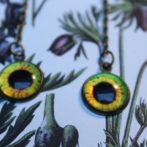 Peut inclure: Une paire de boucles d'oreilles en bronze avec des pendentifs ronds en forme d'œil en verre. Chaque pendentif a une pupille noire, entourée d'un iris jaune et vert avec des accents rouges. Les boucles d'oreilles sont suspendues à de fines chaînes.