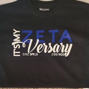 以下が含まれることがあります： 白い文字で「It's my Zeta Versary」と書かれた黒いTシャツ。下に「EOZ SPR 21」と「ZΦB 1920」の文字があります。