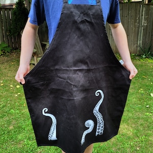 Peut inclure: Tablier noir avec un motif de pieuvre blanc. La pieuvre a trois tentacules avec des ventouses.