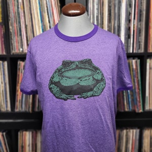 Puede incluir: Camiseta morada con un diseño de rana verde y negra. La camiseta tiene cuello y ribetes de manga morados. La camiseta se muestra en un maniquí. El fondo muestra una colección de discos de vinilo.