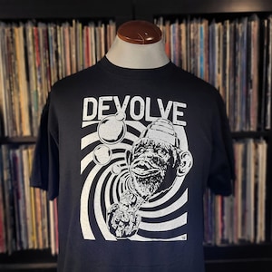 Może przedstawiać: Czarny T-shirt z białym nadrukiem przedstawiającym małpę w kapeluszu dmuchającą bańki. Małpa znajduje się przed wirującym czarno-białym wzorem. Tekst "DEVOLVE" jest nadrukowany nad małpą.