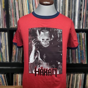 May include: Red ringer t-shirt with a dark navy blue collar and sleeve trim. The shirt features a black and white graphic of a creature with the word "HÄXAN" in red gothic lettering. The shirt is displayed on a mannequin.