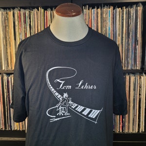 Puede incluir: Camiseta negra con un gráfico blanco de un hombre tocando un piano con el texto "Tom Lehrer" encima.