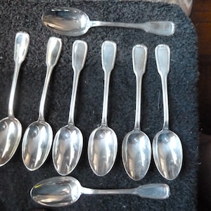 May include: A set of nine silver spoons with ornate handles. The spoons are arranged in a row on a dark surface.