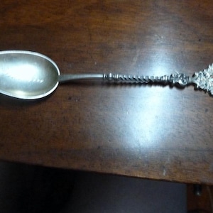 May include: A silver spoon with a decorative handle featuring a cross and floral motifs. The spoon has a rounded bowl and a long, twisted handle.