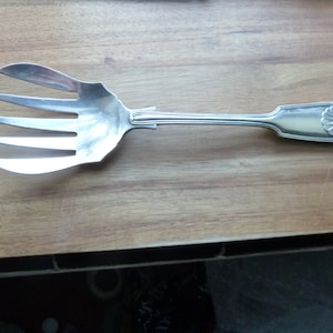 May include: A silver serving fork with four tines and a wide, curved blade. The handle features a shell design. The utensil is placed on a wooden surface.