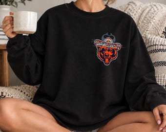 Puede incluir: Una sudadera negra de manga larga y cuello redondo con un gráfico en el pecho izquierdo de un oso que lleva un sombrero de colores. El oso está representado en tonos naranja, azul marino y blanco con la boca abierta mostrando dientes.