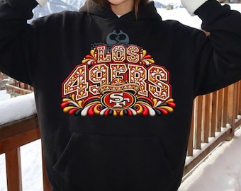 Puede incluir: Una sudadera con capucha negra que presenta un estampado gráfico con el texto LOS 49ERS en una fuente decorativa. El diseño incluye un logotipo central de los San Francisco 49ers, un sombrero y patrones florales en rojo, oro y blanco.