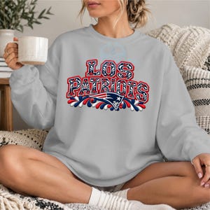 Puede incluir: Una sudadera gris claro de cuello redondo con el texto LOS PATRIOTS en una fuente decorativa llamativa con patrones de filigrana en rojo, blanco y azul. Debajo del texto hay un logotipo estilizado del equipo Patriots con detalles en rojo, blanco y azul.