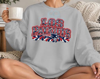 Puede incluir: Una sudadera gris claro de cuello redondo con el texto LOS PATRIOTS en una fuente decorativa llamativa con patrones de filigrana en rojo, blanco y azul. Debajo del texto hay un logotipo estilizado del equipo Patriots con detalles en rojo, blanco y azul.