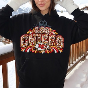 Puede incluir: Una sudadera negra con capucha que presenta un estampado gráfico en el pecho con el texto LOS CHIEFS en letras negritas y ornamentadas. El diseño incluye adornos decorativos en rojo, dorado y blanco y un emblema central.