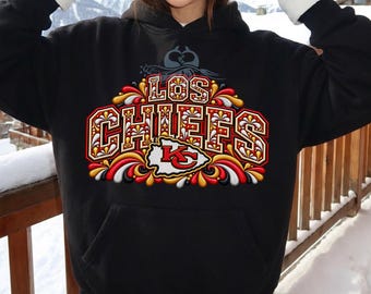 Puede incluir: Una sudadera negra con capucha que presenta un estampado gráfico en el pecho con el texto LOS CHIEFS en letras negritas y ornamentadas. El diseño incluye adornos decorativos en rojo, dorado y blanco y un emblema central.