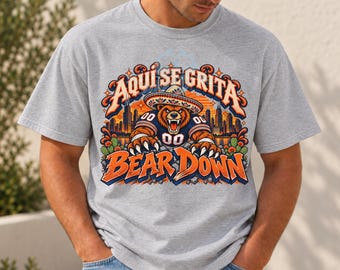 Puede incluir: Una camiseta gris jaspeado con un gráfico de un oso gruñendo que lleva un sombrero. El texto "AQUI SE GRITA" y "BEAR DOWN" aparece en letras naranjas y azules, con un horizonte urbano y siluetas de cactus de fondo.