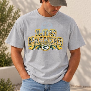 Puede incluir: Una camiseta gris jaspeado de manga corta con el texto LOS PACKERS en una fuente decorativa amarilla, verde y blanca. Debajo del texto hay un logotipo G verde y amarillo rodeado de motivos florales y hojas estilizadas.