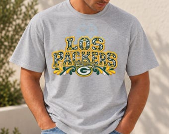 Puede incluir: Una camiseta gris jaspeado de manga corta con el texto LOS PACKERS en una fuente decorativa amarilla, verde y blanca. Debajo del texto hay un logotipo G verde y amarillo rodeado de motivos florales y hojas estilizadas.