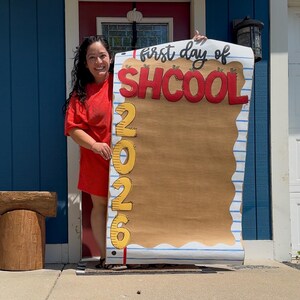 Puede incluir: Un letrero rectangular grande dise&ntilde;ado para parecer una hoja de papel rayado con un centro color canela. El letrero presenta el texto "first day of SHCOOL" en negro y rojo, con el a&ntilde;o vertical "2026" en amarillo en el lado izquierdo.