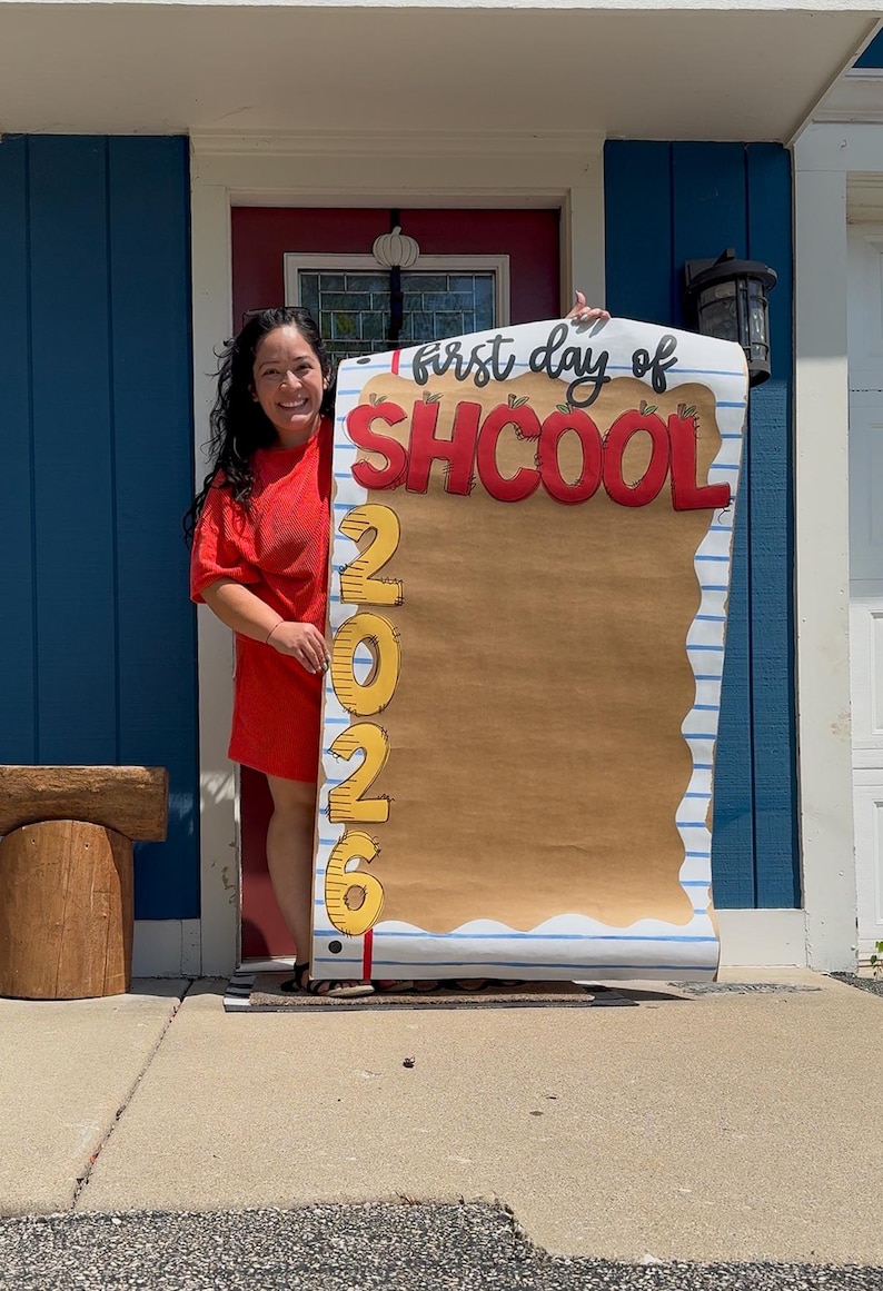 Puede incluir: Un letrero rectangular grande dise&ntilde;ado para parecer una hoja de papel rayado con un centro color canela. El letrero presenta el texto "first day of SHCOOL" en negro y rojo, con el a&ntilde;o vertical "2026" en amarillo en el lado izquierdo.