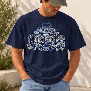Puede incluir: Una camiseta de manga corta azul marino con un estampado gráfico que muestra el texto LOS COWBOYS en letras de bloque blancas y azules. El diseño incluye remolinos decorativos, estrellas y un emblema central de estrella.