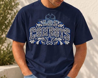 Puede incluir: Una camiseta de manga corta azul marino con un estampado gráfico que muestra el texto LOS COWBOYS en letras de bloque blancas y azules. El diseño incluye remolinos decorativos, estrellas y un emblema central de estrella.