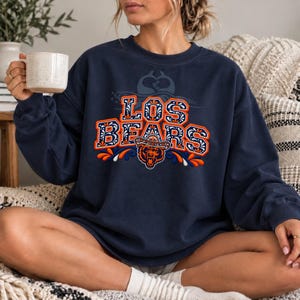 Puede incluir: Una sudadera azul marino de cuello redondo con el texto LOS BEARS en una fuente decorativa naranja y blanca. Debajo del texto hay un gráfico de un oso con sombrero, acentuado con patrones florales naranjas y azules y formas de hojas.