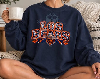 Puede incluir: Una sudadera azul marino de cuello redondo con el texto LOS BEARS en una fuente decorativa naranja y blanca. Debajo del texto hay un gráfico de un oso con sombrero, acentuado con patrones florales naranjas y azules y formas de hojas.
