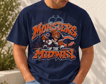 Puede incluir: Una camiseta azul marino de manga corta con un gráfico de un oso pardo con uniforme de fútbol americano. El texto dice Monsters of the Midway en letras naranjas y blancas, con un horizonte urbano estilizado y una gran letra C naranja.