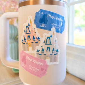 Puede incluir: Un vaso térmico blanco con asa, con dos pegatinas estilo billete "Magic Kingdom". Las pegatinas representan un castillo y dicen "EST. 1971" y "ADMIT ONE". El vaso tiene una tapa transparente y un borde dorado.