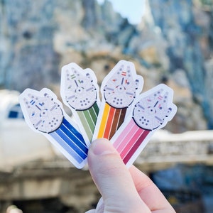 May include: Four colorful stickers featuring the Millennium Falcon starship. Each sticker has a different colored stripe design, including blue, green, orange, and pink. The stickers are held in front of a blurred background of a Star Wars-themed landscape.