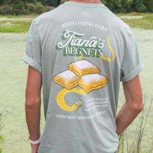 May include: A light green t-shirt with a graphic of a cartoon bee and beignets. The text on the shirt reads "Baked Fresh Daily Tiana's Beignets", "Powdered Sugar 15¢", "Cinnamon Sugar 15¢", "Wild Honey 20¢", and "Voted Best Beignets in T-Town".