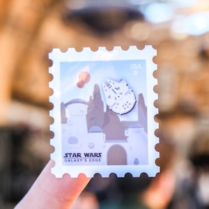 May include: A white and blue Star Wars Galaxy's Edge commemorative postage stamp featuring the Millennium Falcon flying over a desert landscape. The stamp is printed with "USA" and "Star Wars Galaxy's Edge".