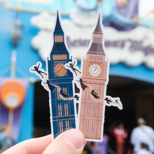 May include: Two paper cutouts of Big Ben, one blue and one brown, with silhouettes of Peter Pan and the Darling children flying around them. The cutouts are held in front of a blue and purple background.